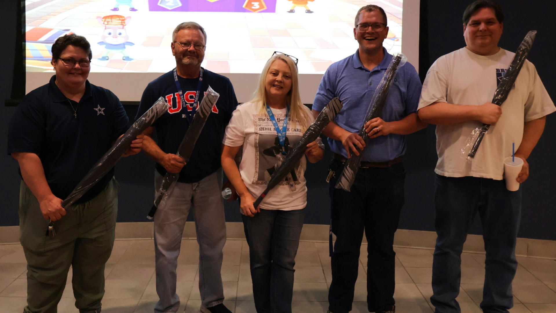 Five smiling faculty members stand on stage each holding a wrapped prize, with a colorful game show displayed on the screen behind them.